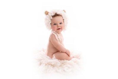 A baby in a furry hat sitting on a white background.