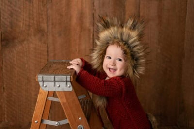A baby wearing a furry hat on a ladder.