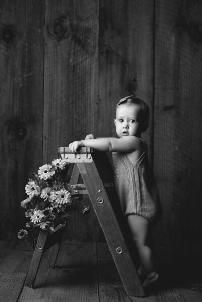 A black and white photo of a baby leaning on a wooden ladder.