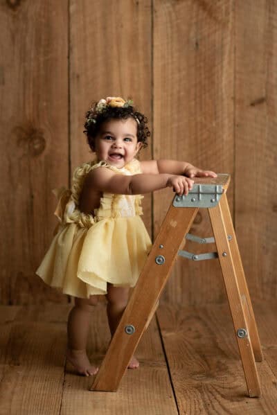 A baby girl in a yellow dress is standing on a wooden ladder.