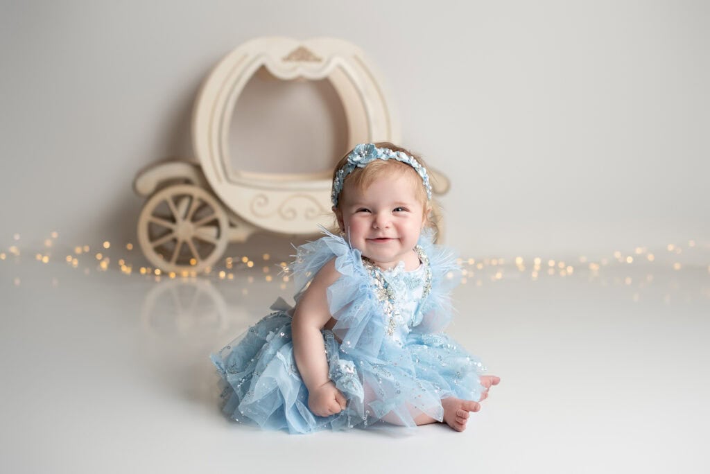 A smiling baby in a frilly blue dress sits on the floor, capturing the essence of baby photography. A decorative wooden carriage is in the background with soft, glowing string lights draped around. Tiny Dreamers Studio - Maternity & Newborn Photographer.