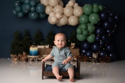 A baby sitting on a wooden chair in front of balloons.