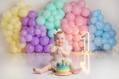 A baby girl sitting in front of a rainbow cake with balloons.