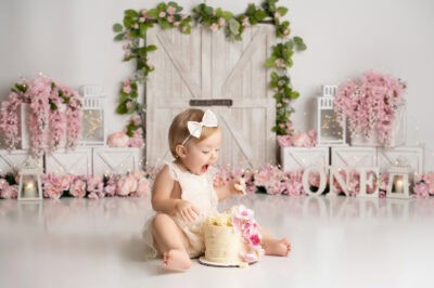 A baby girl is eating a cake in front of a pink backdrop.
