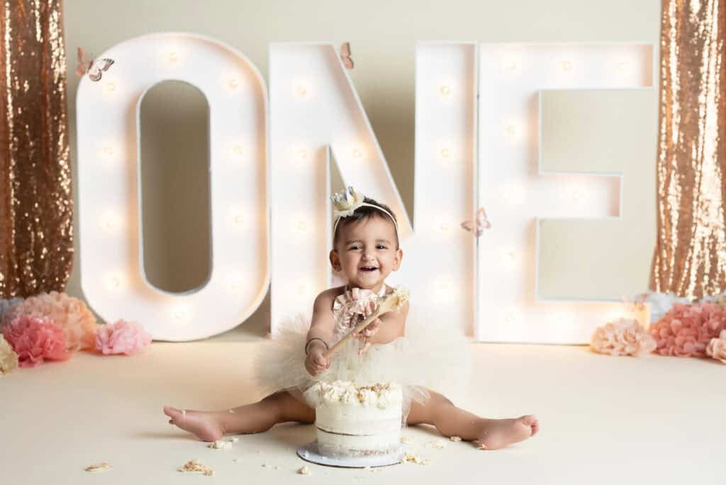 A baby girl sitting in front of a cake with the number one in front of it.