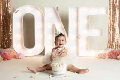 A baby girl sitting in front of a cake with the number one in front of it.