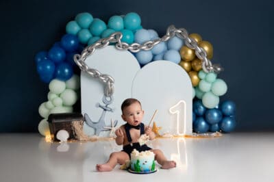 A baby sitting in front of a cake and balloons.