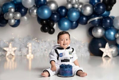 A baby in an astronaut costume sitting in front of balloons.