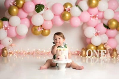 A baby girl sitting in front of a pink and gold balloon backdrop.