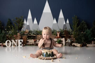 A baby boy eating a cake in front of a forest.