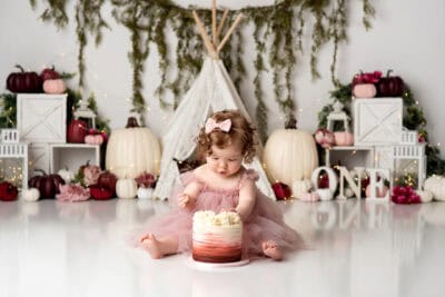 A little girl in a pink dress eating a cake in front of pumpkins and teepees.