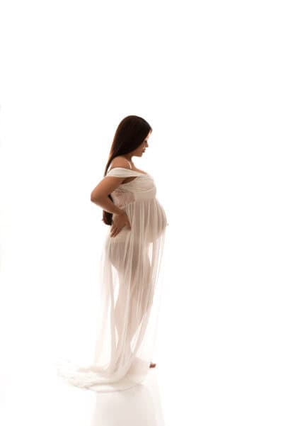 A pregnant woman in a white dress posing in front of a white background.