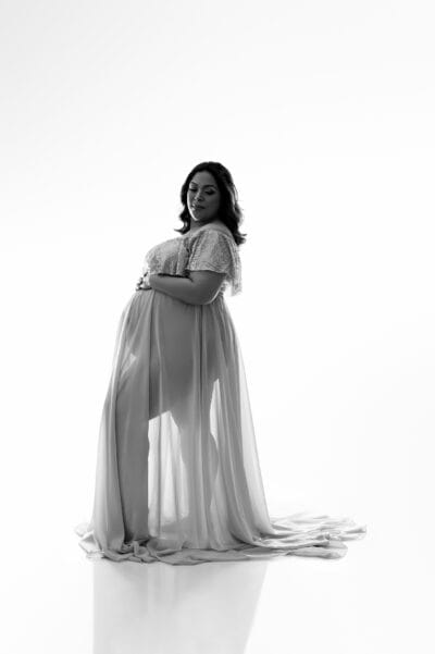 A pregnant woman in a white dress posing in front of a white background.