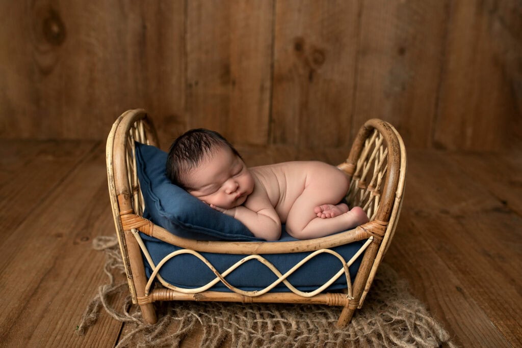 In this serene example of newborn photography, a baby sleeps peacefully on a small rattan bed with a navy blue cushion. The bed rests on rustic wooden floors, complemented by a burlap cloth. The wooden wall in the background adds to the cozy and warm ambiance. Tiny Dreamers Studio - Maternity & Newborn Photographer.