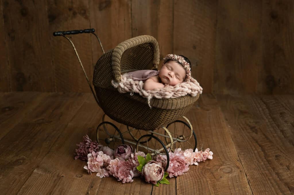 A newborn sleeps peacefully in a vintage wicker pram adorned with a soft blanket and floral headband. The scene, perfect for photography, is set against a rustic wooden background, surrounded by pink and mauve flowers. Tiny Dreamers Studio - Maternity & Newborn Photographer.