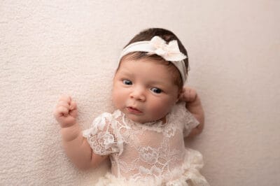 A baby girl in a white dress posing against a wall.