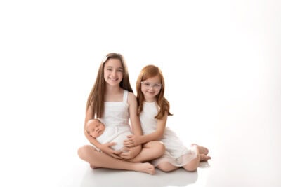 Three little girls sitting on a white background holding a baby.