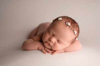 A baby girl wearing a flower headband is laying on a white background.