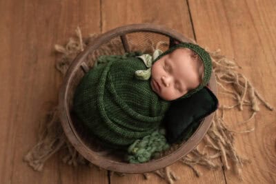 A baby sleeping in a green knitted blanket in a wooden basket.