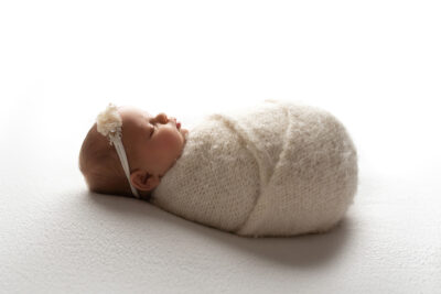 A newborn wrapped in a white blanket on a white background.