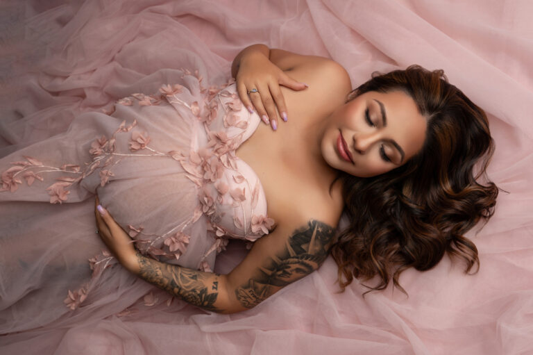 a pregnant woman lies on a pink, flowing fabric. she has long brown hair and wears a strapless, flower adorned, pink gown. her left arm rests on her belly, and her right arm, showing detailed tattoos, is placed above her. this maternity photography captures her serene expression with closed eyes beautifully.