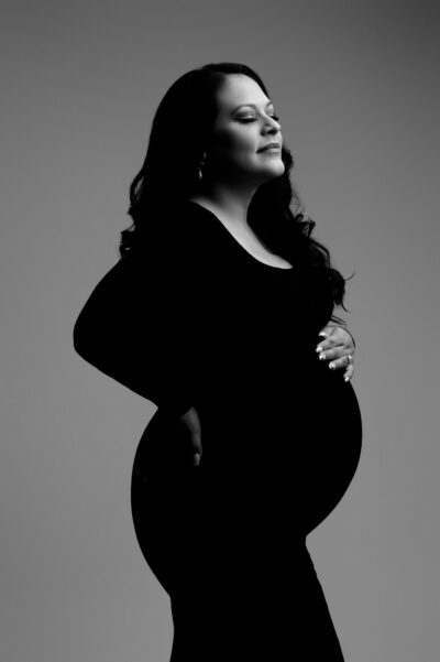 a pregnant woman in a side profile position, wearing a form fitting black dress and cradling her belly, poses for a maternity photography session against a grey background with soft lighting.