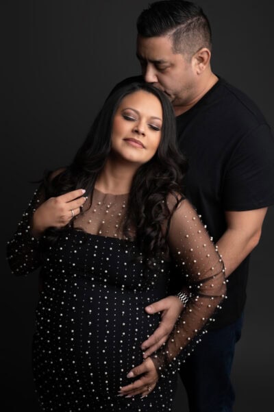 a pregnant woman in a beaded black dress stands with her eyes closed while a man stands behind her, embracing her and touching her belly, against a dark background during a maternity photo session.