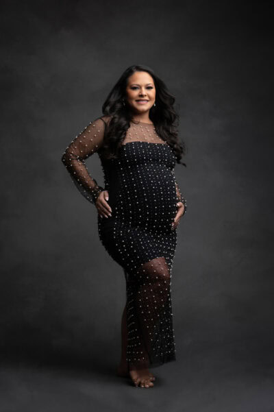 a confident woman wearing a sheer, black, beaded gown stands with one hand on her hip against a dark gray background during her maternity photo session. she has long dark hair and a radiant smile.