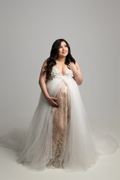 a radiant pregnant woman in an elegant white and gold gown, smiling gently, stands confidently against a neutral background for her maternity photo session. her dress is sheer and embellished with floral patterns.