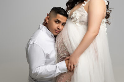 a man in a white shirt embraces a pregnant woman in a white and beige maternity dress, both posing tenderly against a light gray background during their maternity photo session.