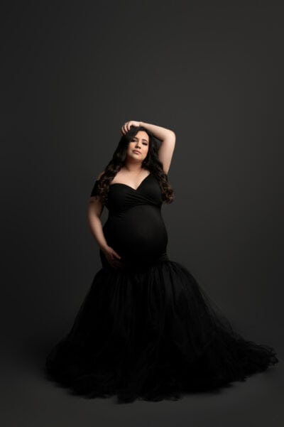 a pregnant woman in a flowing black dress stands gracefully against a dark background, her hand on her forehead looking thoughtful during her maternity photo session.