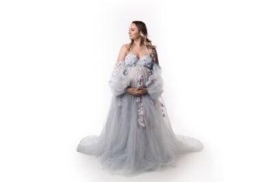a pregnant woman in an elegant, flowing pale blue gown cradling her belly, standing against a white background, exuding a serene expression during her maternity photo session.
