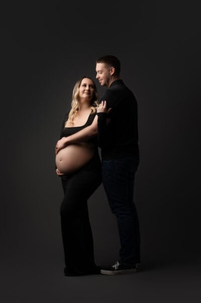 a pregnant woman and a man stand together in a maternity photography session, the man behind her touching her belly. they are smiling, dressed in black, set against a dark background.