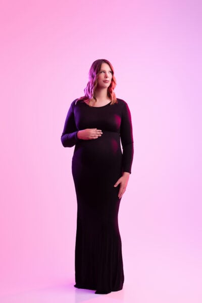 a pregnant woman in a long black dress stands gracefully, touching her belly, against a soft pink background, looking gently towards the side during her maternity photo session.