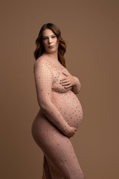 pregnant woman in a sheer, embellished gown stands gracefully for her maternity photography session, her hands gently cradling her belly against a soft brown background. her expression is serene and contemplative