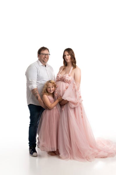 a maternity photo session featuring a pregnant woman in a flowing pink dress, a young girl in a matching outfit, and a man in a white shirt and jeans, all smiling, against a white background.