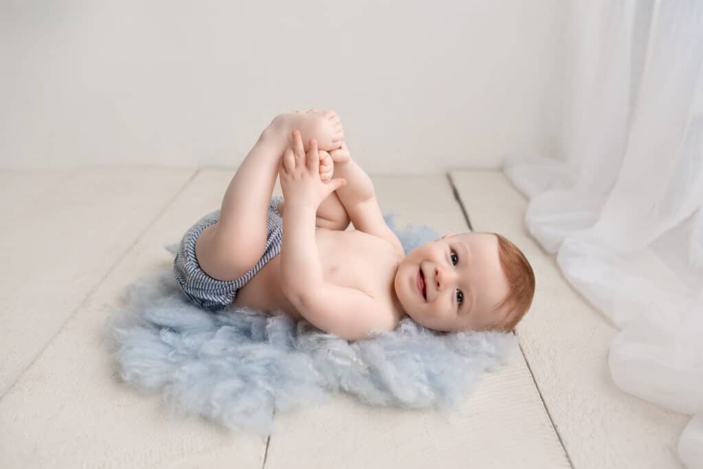 A smiling baby lies on their back on a fluffy blue rug. They are wearing striped shorts and holding their feet with their hands. The setting is a bright room with light wooden floors and sheer white curtains. Tiny Dreamers Studio - Maternity & Newborn Photographer.