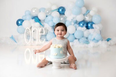 A baby boy sitting in front of a blue cake with balloons.