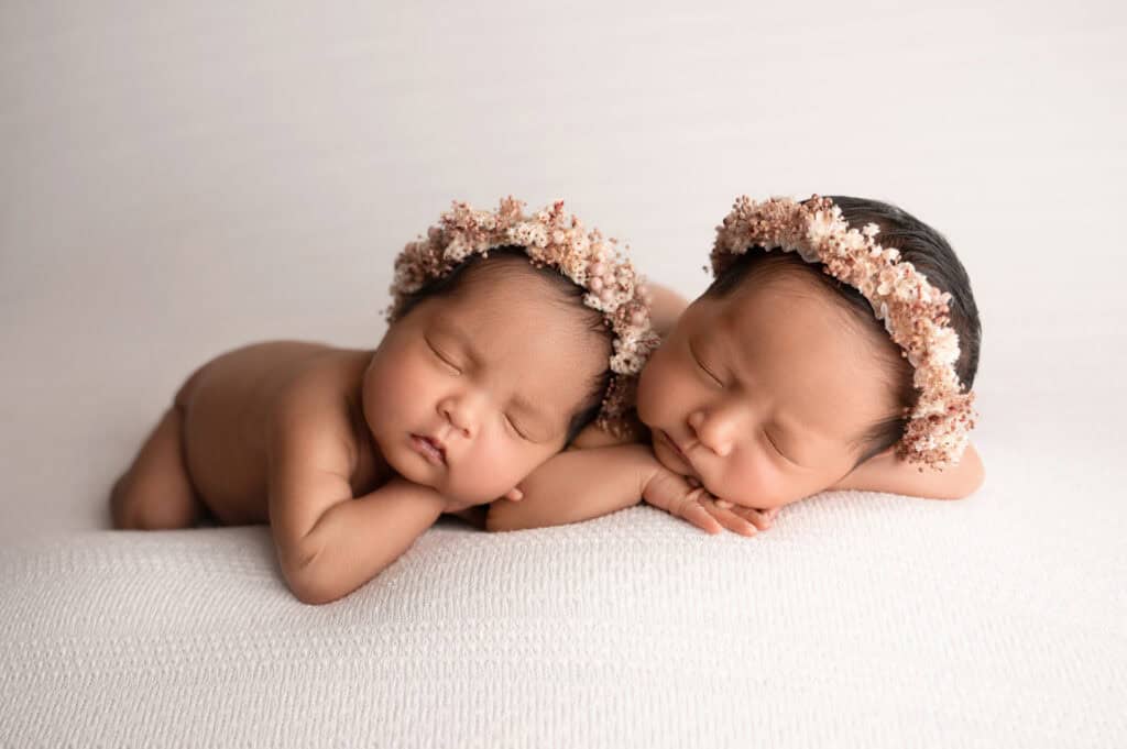 Two sleeping babies lie on a soft white surface, both adorned with delicate floral headbands. Their peaceful expressions convey serenity and innocence. Tiny Dreamers Studio - Maternity & Newborn Photographer.