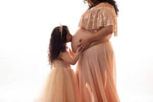 A pregnant woman in a pink dress kisses her daughter's belly.