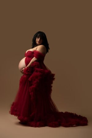 A pregnant woman in a red dress posing on a brown background.