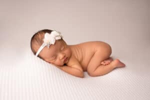 A newborn girl wearing a white headband is laying on a white background.