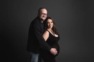 A pregnant couple posing for a photo in front of a black background.