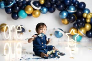A baby is eating cake in front of balloons.