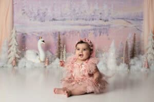 A baby girl in a pink tutu sitting in front of a snowy backdrop.