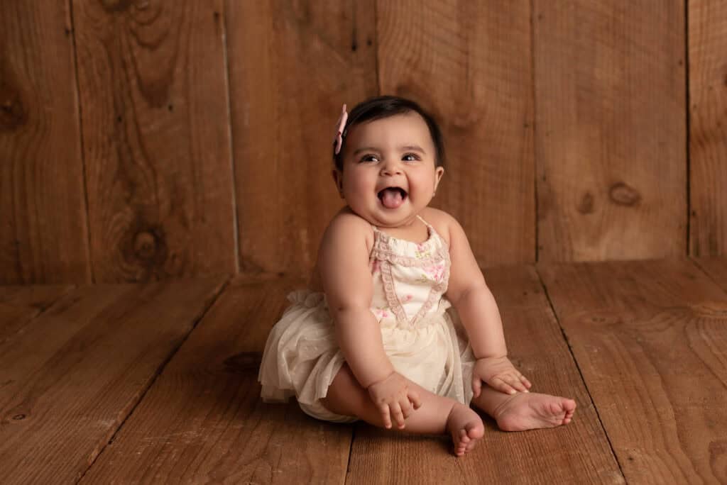 A baby sits on a wooden floor, smiling and sticking its tongue out. The baby is wearing a light-colored dress with pink accents and has a small bow in the hair. The wooden background adds a rustic feel to the scene. Tiny Dreamers Studio - Maternity & Newborn Photographer.