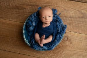 A baby boy in a blue wrap sitting in a bowl on a wooden floor.
