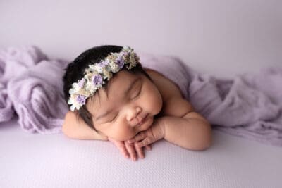 A baby girl sleeping on a purple blanket with a flower crown on her head.