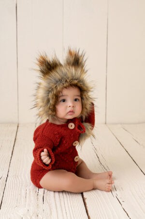 A baby wearing a red furry hat sitting on a wooden floor.