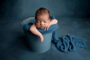 A baby boy sleeping in a bucket on a blue blanket.
